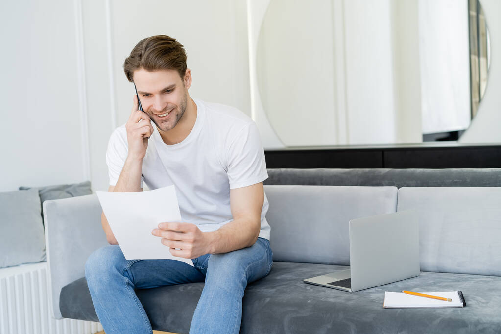pozitív férfi beszél mobilon, miközben nézi dokumentum közelében laptop kanapén - Fotó, kép
