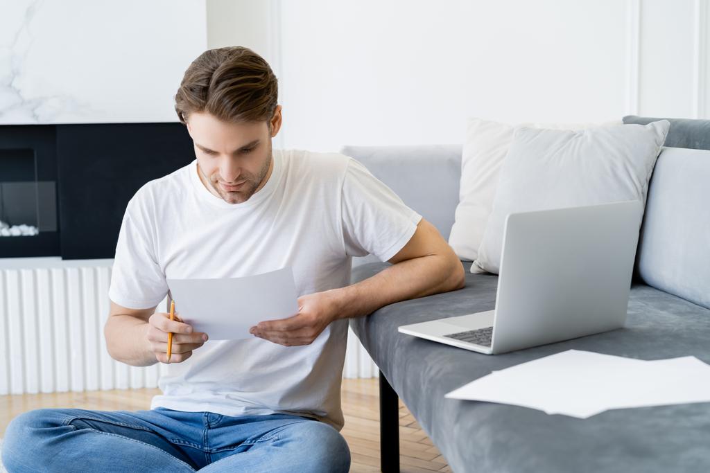 мужчина смотрит на документ, сидя рядом с ноутбуком, и бумаги на диване - Фото, изображение