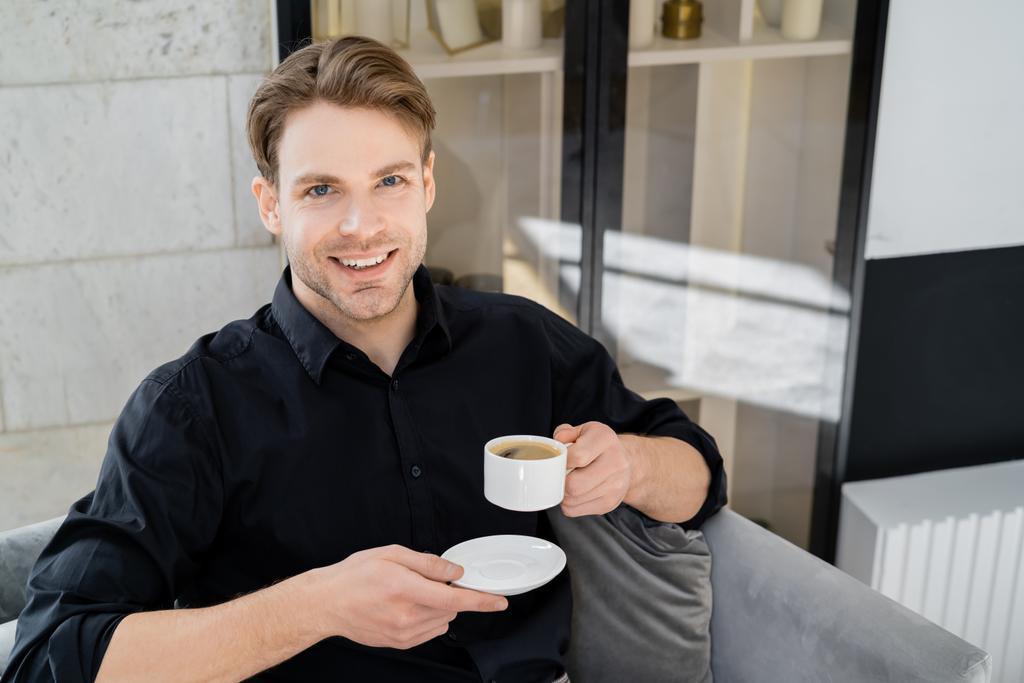 χαρούμενος άνθρωπος με φλιτζάνι καφέ και πιατάκι κοιτάζοντας κάμερα, ενώ κάθεται στο σπίτι - Φωτογραφία, εικόνα