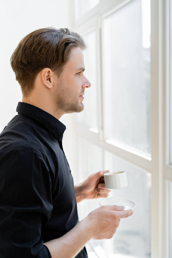vista lateral del hombre en camisa negra de pie con taza de café cerca de la ventana - Foto, imagen
