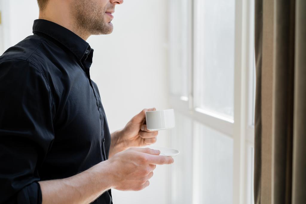 Siyah gömlekli, elinde fincan tabağı ve pencerenin yanında kahve fincanı olan kırpık bir adamın yan görüntüsü. - Fotoğraf, Görsel