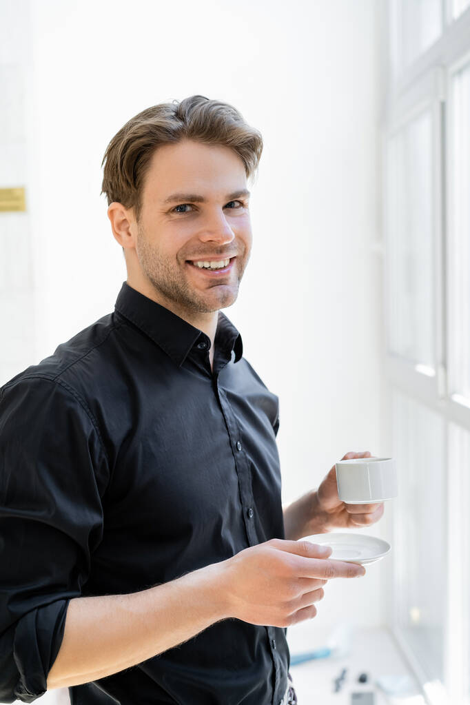 счастливый человек в черной рубашке держит чашку кофе и улыбается в камеру - Фото, изображение