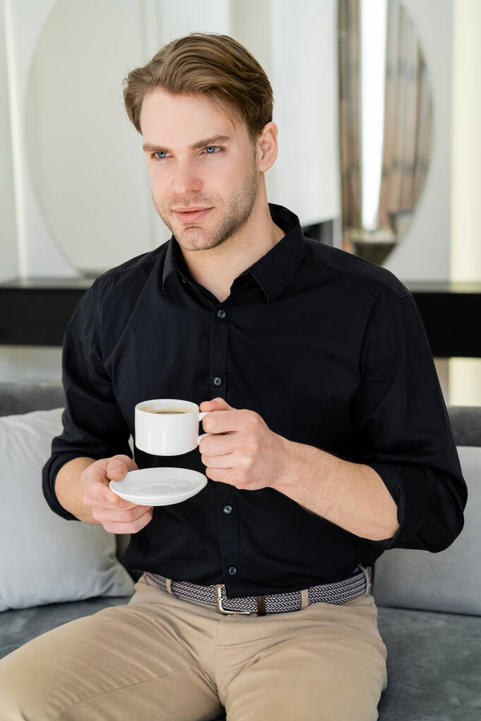 homem de camisa preta sentado com xícara de café e pires no sofá na sala de estar  - Foto, Imagem
