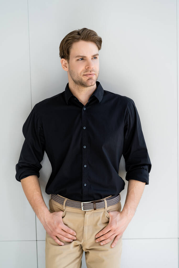 стильний чоловік у чорній сорочці та бежевих брюках, що стоять з великими пальцями в кишенях біля сірої стіни
 - Фото, зображення