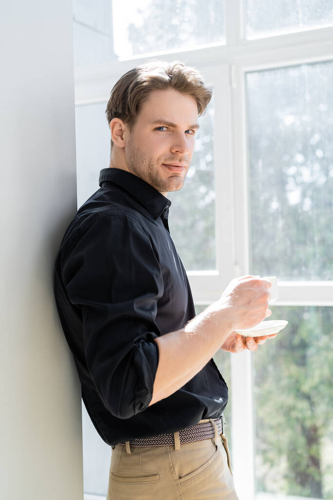 молодий чоловік стоїть біля вікна з чашкою кави і дивиться на камеру
 - Фото, зображення