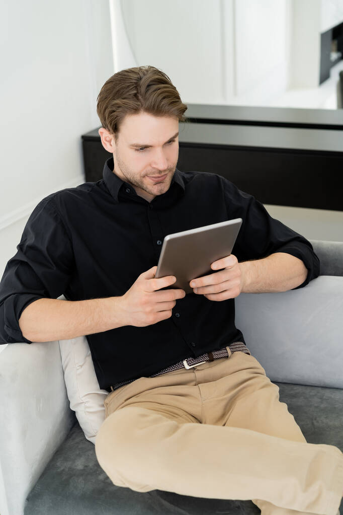 fiatal férfi fekete ingben ül a kanapén otthon, és használja a digitális tabletta - Fotó, kép