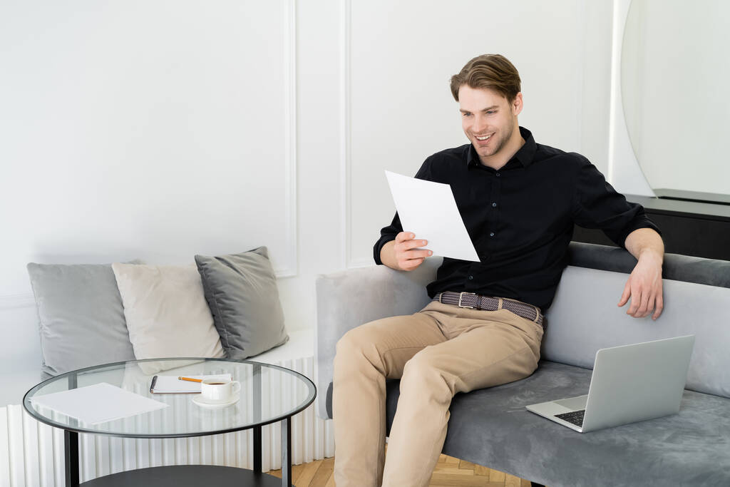 веселый человек читает документ, сидя на диване возле ноутбука и журнального столика - Фото, изображение