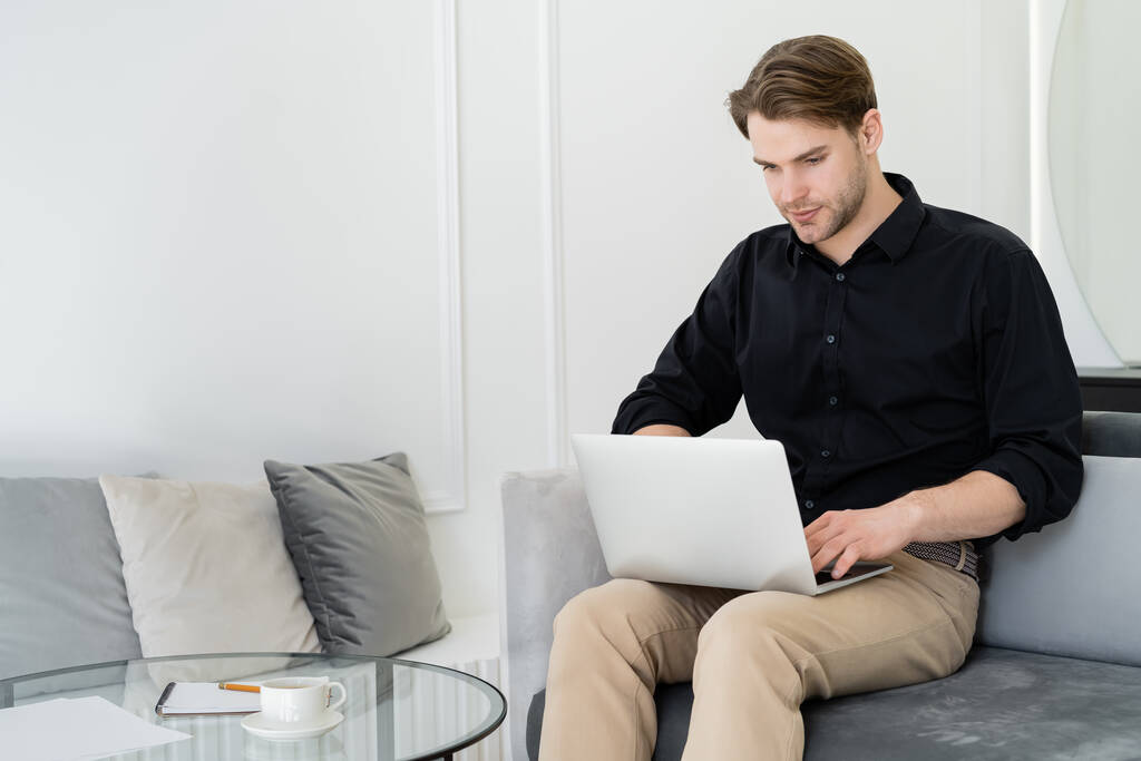 человек работает на ноутбуке рядом с журнальным столиком с чашкой и ноутбуком - Фото, изображение