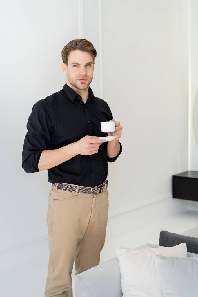 junger Mann in schwarzem Hemd und beiger Hose steht mit Kaffeetasse und Untertasse im Wohnzimmer - Foto, Bild