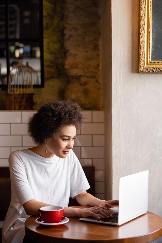 jeune femme afro-américaine en utilisant un ordinateur portable près d'une tasse de café dans un café  - Photo, image