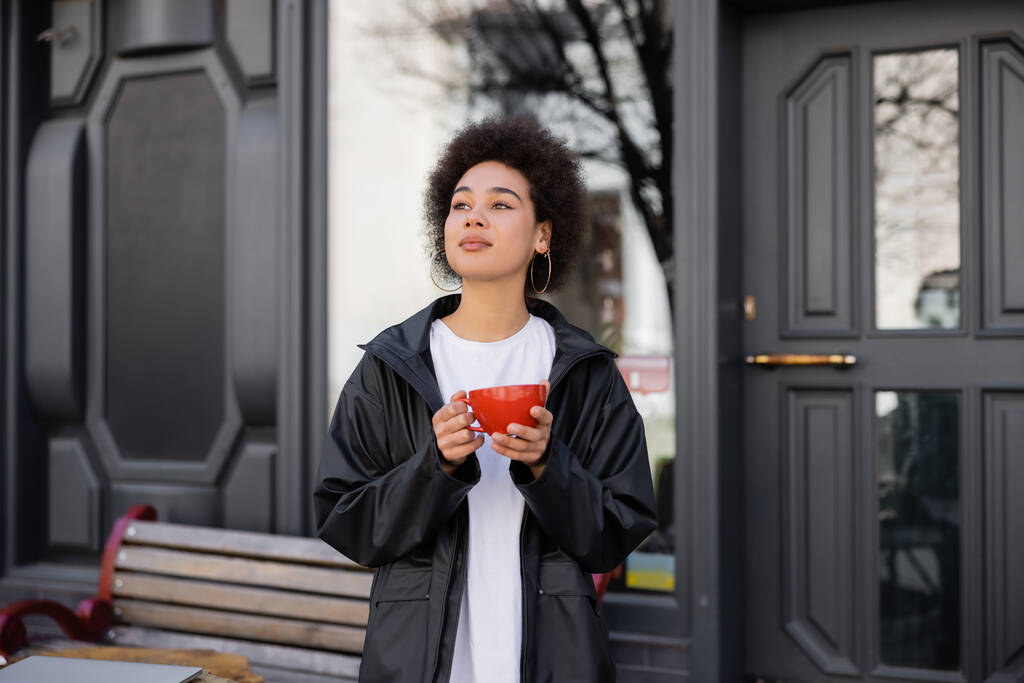 Sokakta elinde kahve tutan ceketli Afro-Amerikan kadın.  - Fotoğraf, Görsel