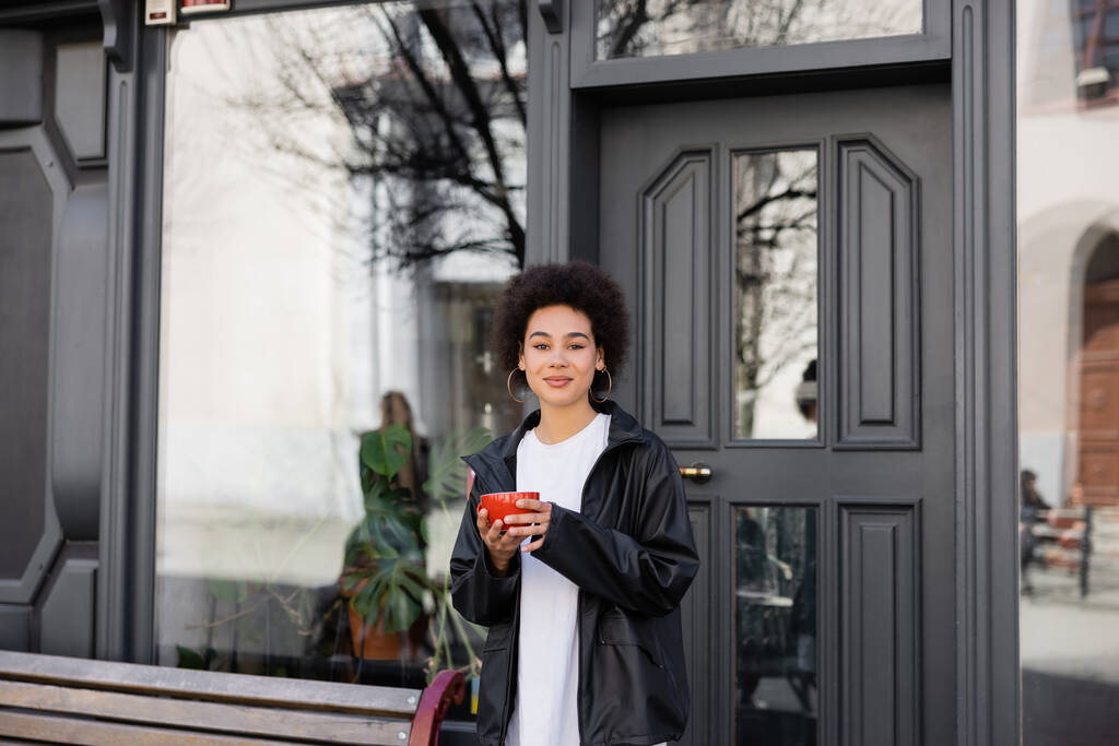 pleased african american woman in jacket holding cup of coffee on street  - Foto, afbeelding