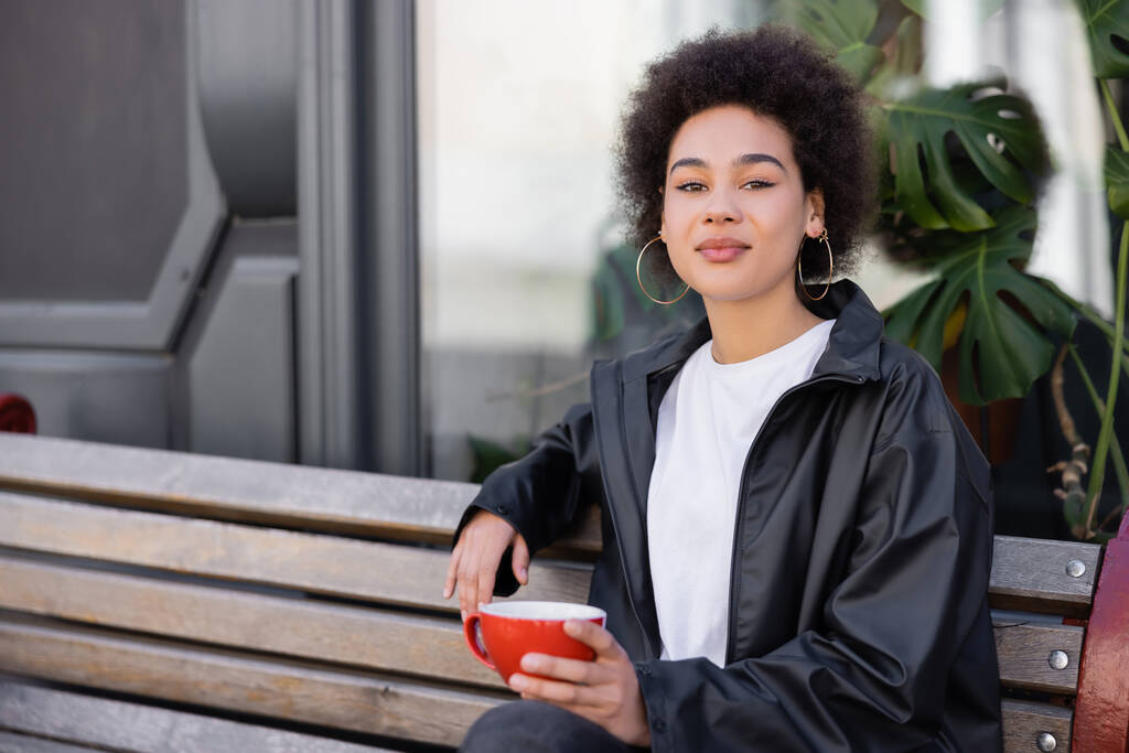 joven afroamericana mujer en chaqueta sosteniendo taza de café y sentado en el banco  - Foto, imagen