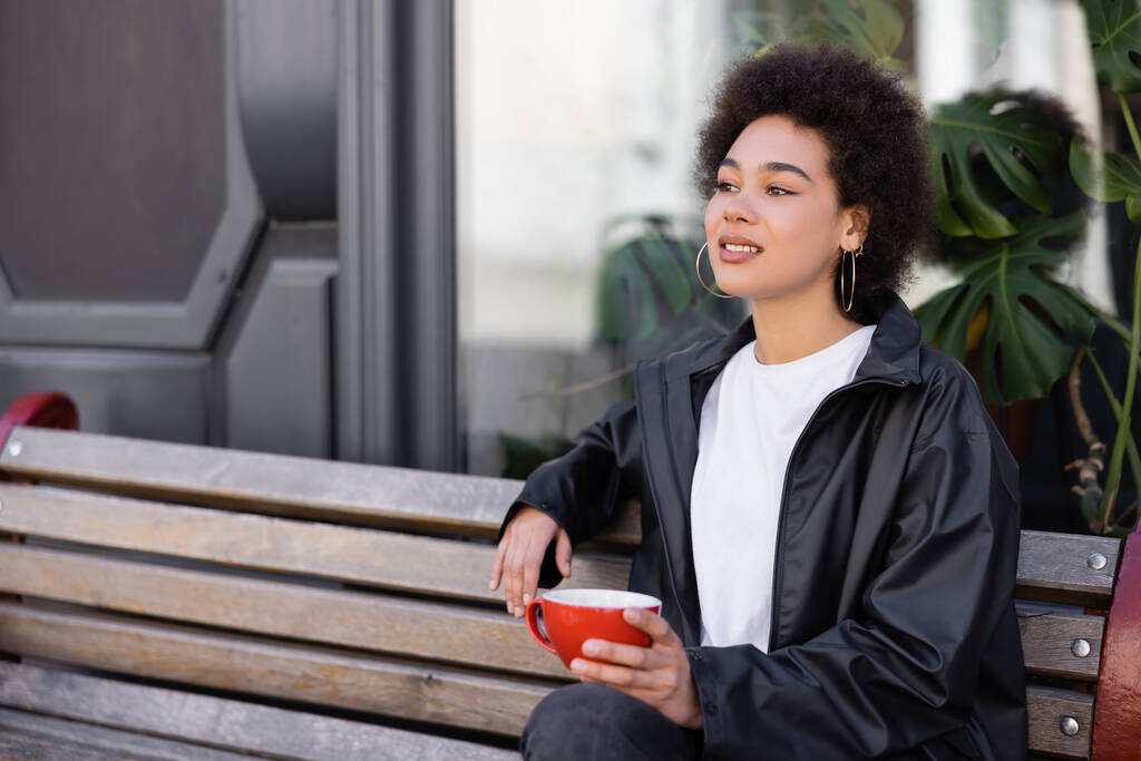 curly african american woman in jacket holding cup of coffee while sitting on bench  - Foto, afbeelding