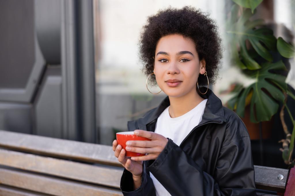 curly african american woman holding cup of coffee while sitting on bench  - Valokuva, kuva