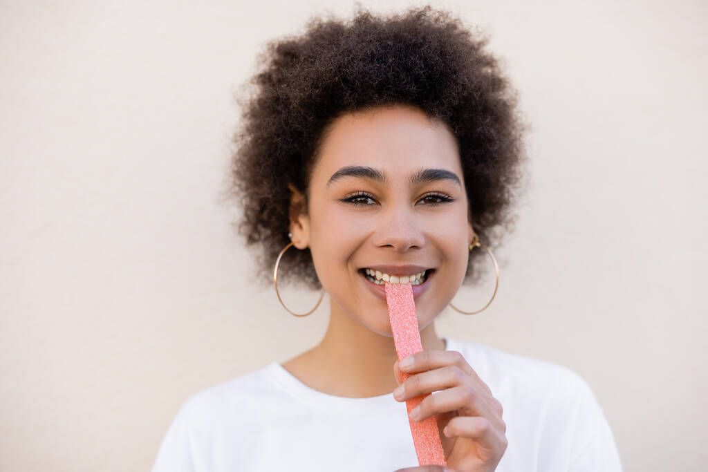 joyful african american young woman eating sweet jelly strip on white - Foto, imagen