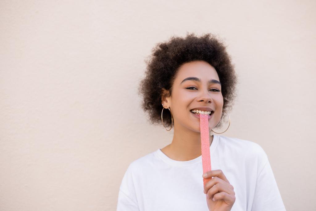 усміхнена афроамериканська молода жінка їсть солодку желе-бульбашкову стрічку на білому
 - Фото, зображення