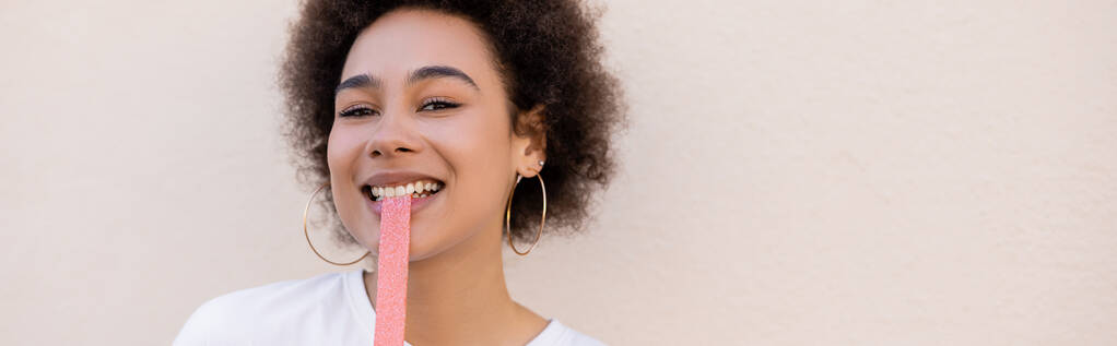 happy african american young woman eating sweet jelly bubble tape on white, banner - Zdjęcie, obraz
