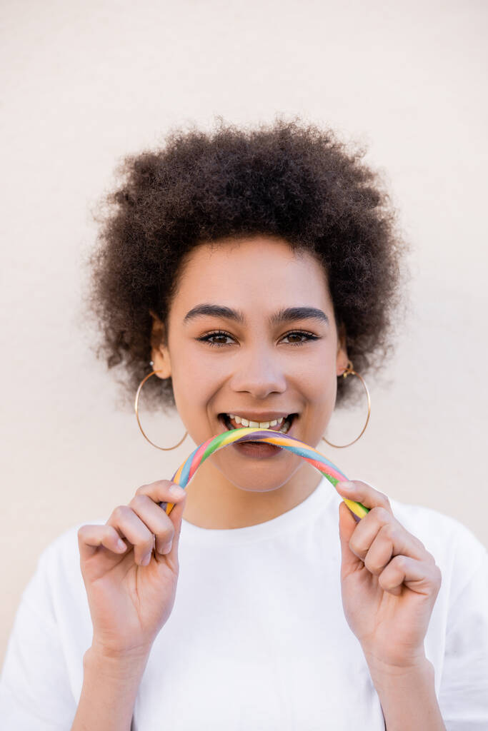 happy african american young woman in hoop earrings eating jelly candy on white - Zdjęcie, obraz