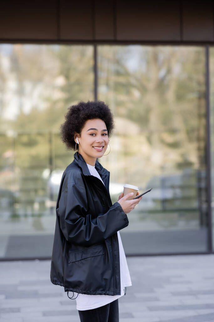 smiling african american woman in earphone holding paper cup and smartphone outside - Foto, Bild