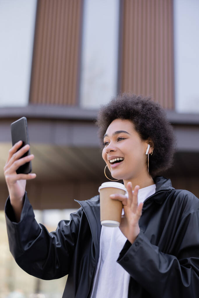 joyful african american woman in earphone holding paper cup and having video chat on smartphone outside - Photo, Image
