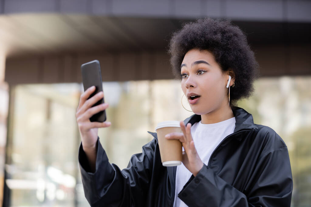 scioccato donna afroamericana in auricolare in possesso di tazza di carta e avendo video chat su smartphone al di fuori - Foto, immagini