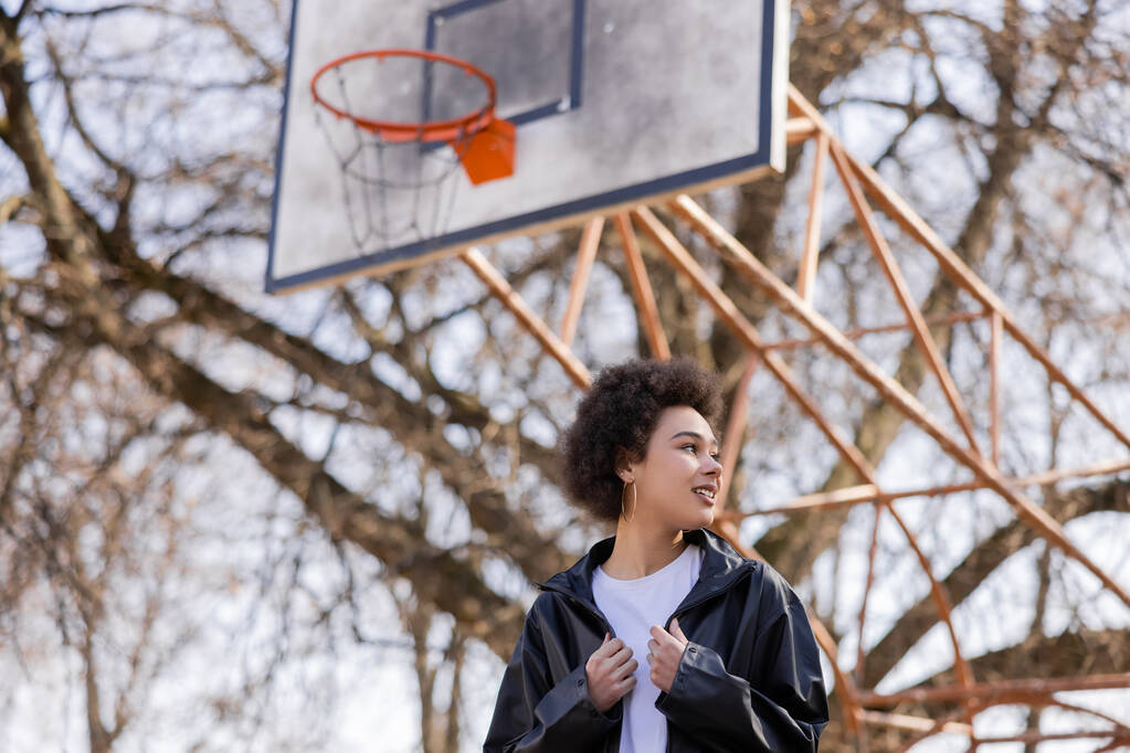 vue à angle bas de femme afro-américaine heureuse en veste debout sur le terrain de basket-ball extérieur  - Photo, image