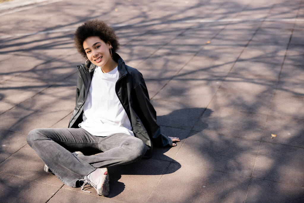 longitud completa de la sonriente mujer afroamericana en chaqueta sentada con las piernas cruzadas sobre asfalto con sombras  - Foto, Imagen