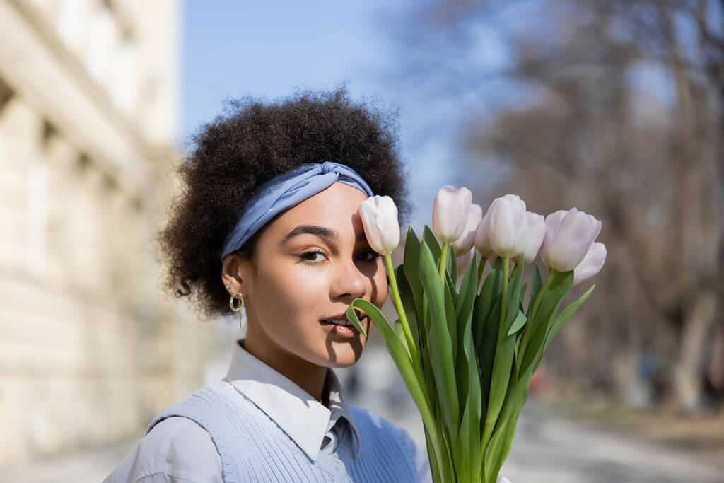 若いアフリカ系アメリカ人女性がチューリップの花束の近くでカメラを見て - 写真・画像