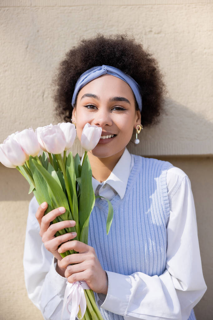 улыбающаяся африканская американка в синем жилете и белой рубашке с букетом тюльпанов у стены - Фото, изображение