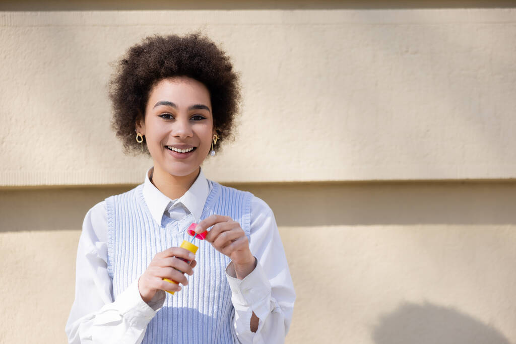 heureuse femme afro-américaine en gilet bleu et chemise blanche tenant bouteille avec bulles de savon - Photo, image