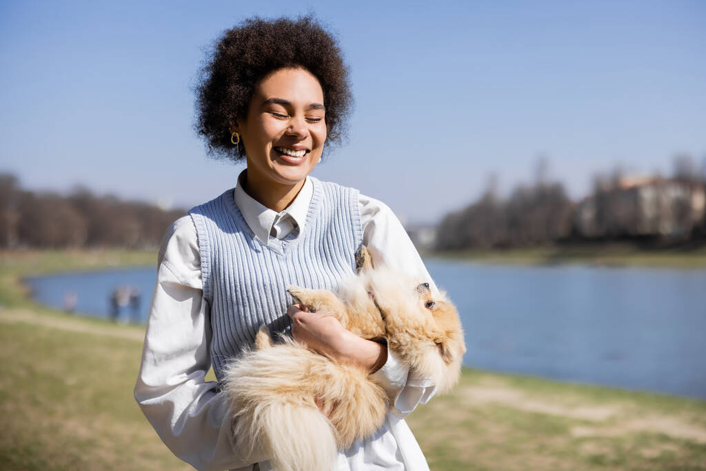 happy african american woman with closed eyes holding pomeranian spitz  - Foto, Imagen
