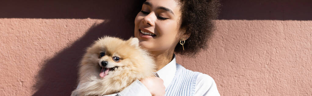happy african american woman in blue vest holding in arms pomeranian spitz, banner - Foto, afbeelding