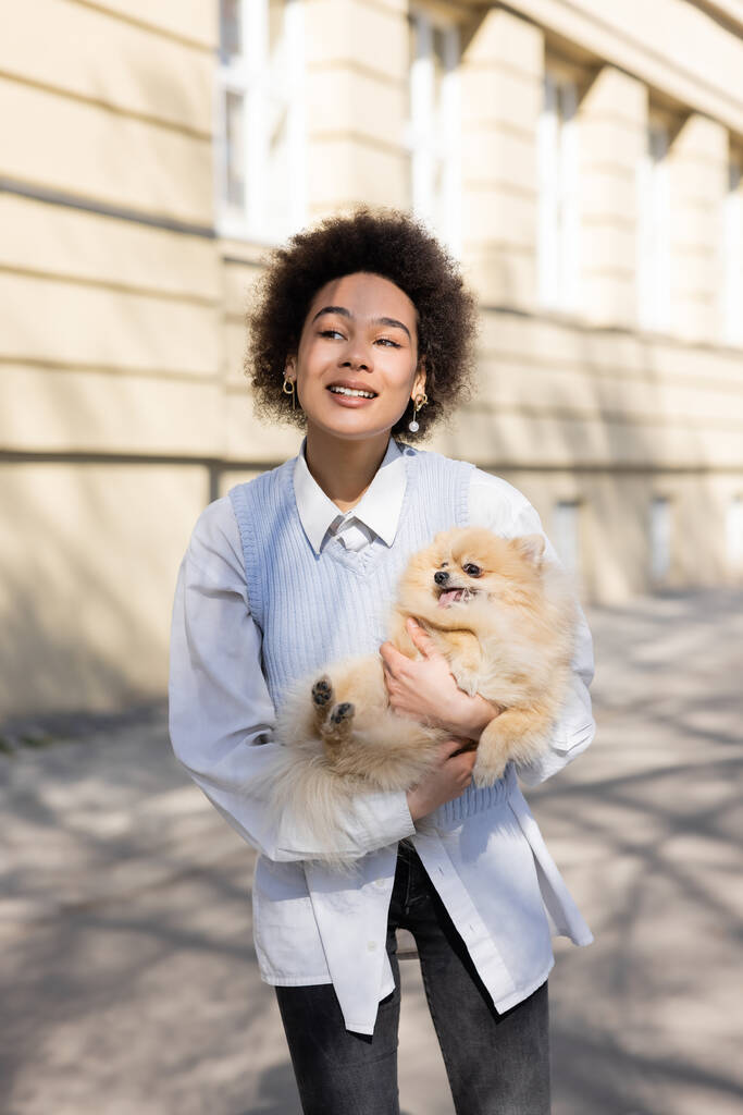 happy african american woman in blue vest holding pomeranian dog on street - Photo, Image