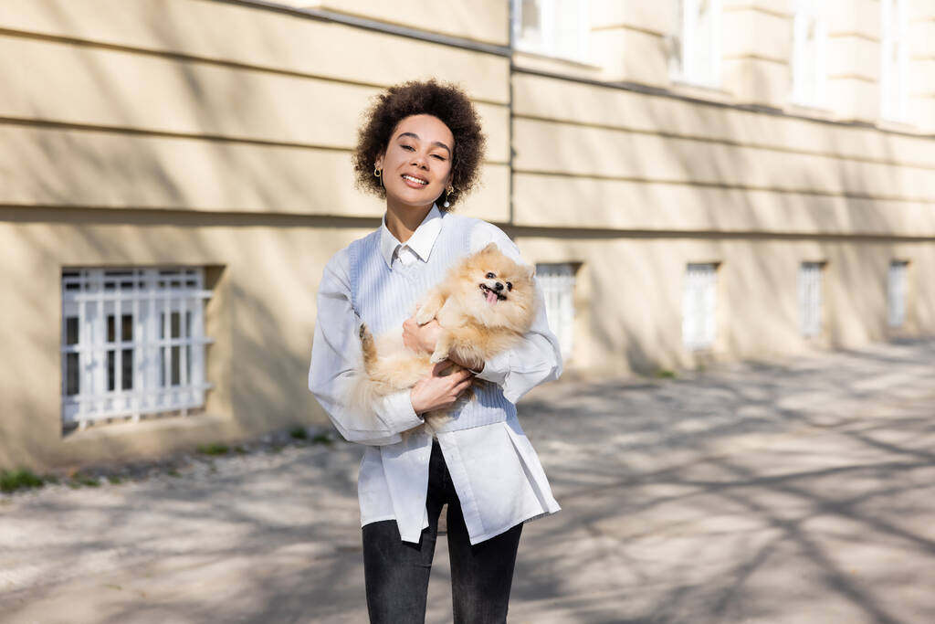positive african american woman in blue vest holding pomeranian dog on street - Foto, imagen