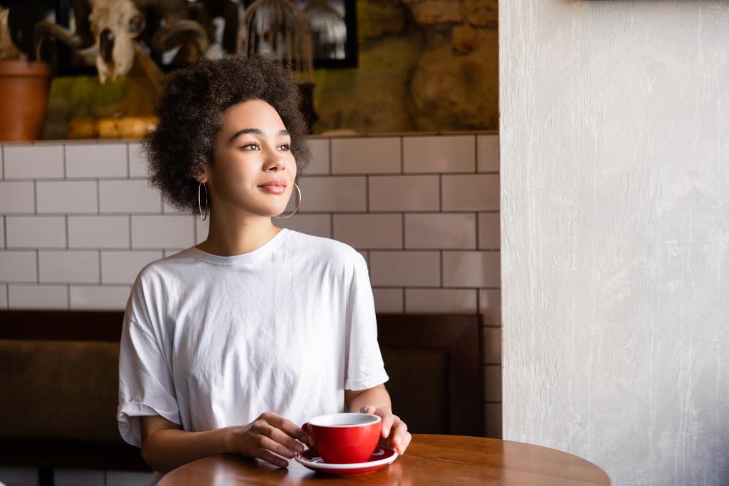 dreamy african american woman in hoop earrings and white t-shirt holding cup - Foto, immagini