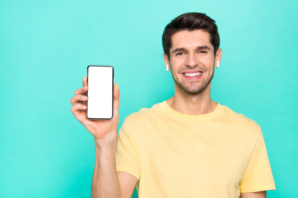 Fotografie legrační brunet mladý chlap show telefon nosí t-shirt sluchátka izolované na pozadí teal color - Fotografie, Obrázek