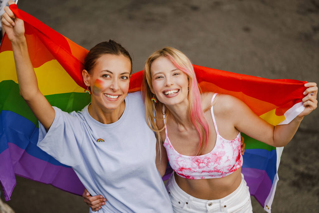 Joven pareja lesbiana caminando con banderas de arco iris durante el desfile de orgullo en la calle de la ciudad - Foto, Imagen
