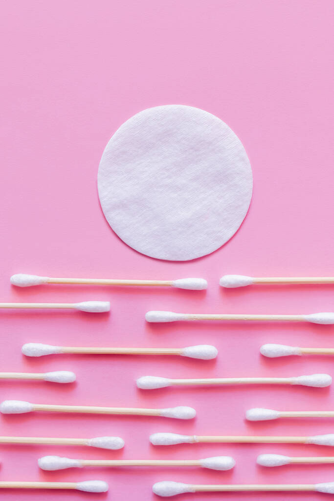 white cotton pad above rows of ear sticks on pink background, top view - Foto, imagen