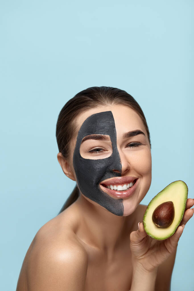 Красивая улыбающаяся женщина в маске на лице, держащая половину авокадо перед лицом. Концепция ухода за кожей красоты. Косметическая маска. Лечение. Косметология - Фото, изображение