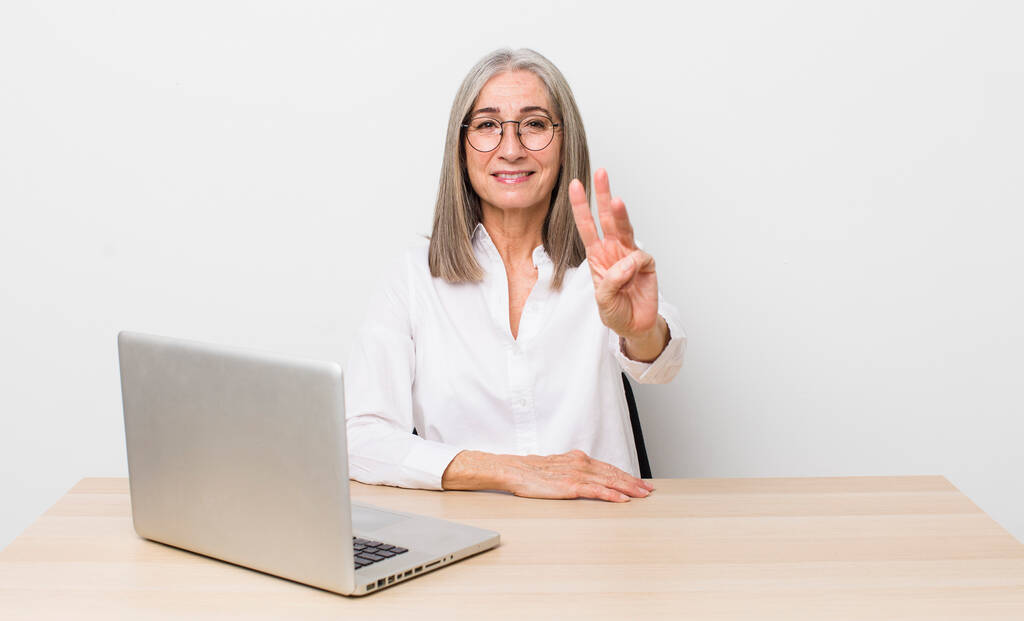 Seniorin mit grauem Haar lächelt freundlich und zeigt Nummer drei. Schreibtisch und Geschäftskonzept - Foto, Bild