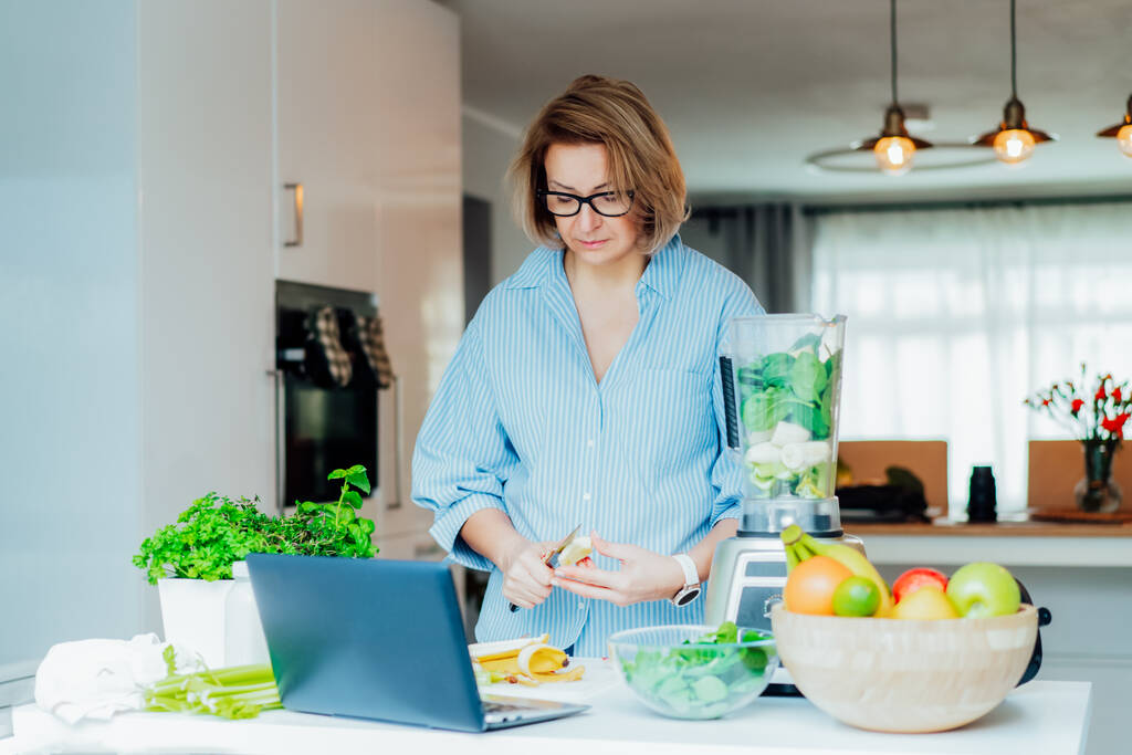 Μεσήλικας γυναίκα βλέποντας ένα μάθημα μαγειρικής βίντεο σε φορητό υπολογιστή, ενώ προετοιμασία ενός γεύματος σε μια κουζίνα. Γυναίκα κάνει πράσινο smoothie. Διαδικτυακή συνταγή. Υγιεινός τρόπος ζωής, πρόγραμμα απώλειας βάρους. - Φωτογραφία, εικόνα