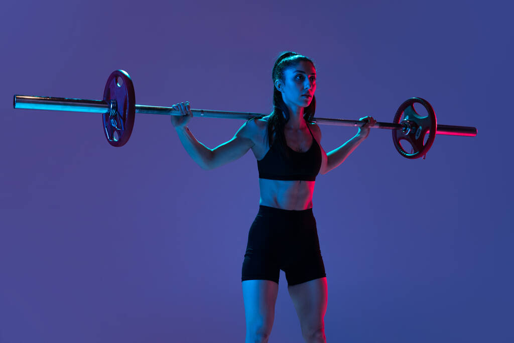 Retrato de mulher musculada em treinamento de sportswear com uma barra isolada em fundo roxo em luz de néon. Esporte, conceito de levantamento de peso - Foto, Imagem
