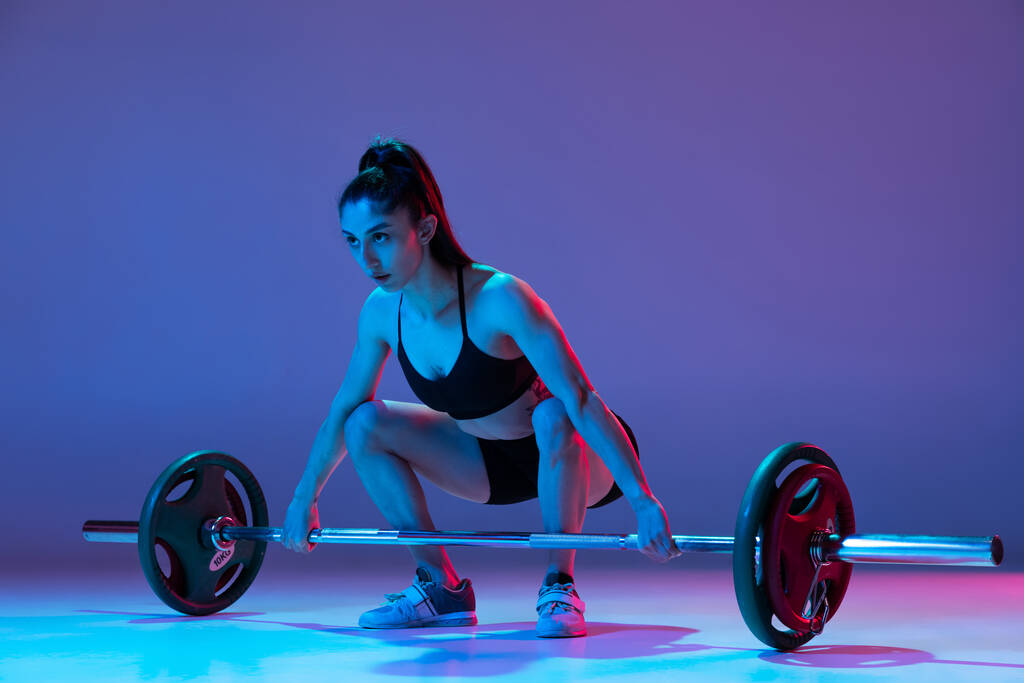 Portret van gespierde vrouw in sportkleding training met een lange halter geïsoleerd op paarse achtergrond in neon licht. Sport, gewichtheffen - Foto, afbeelding