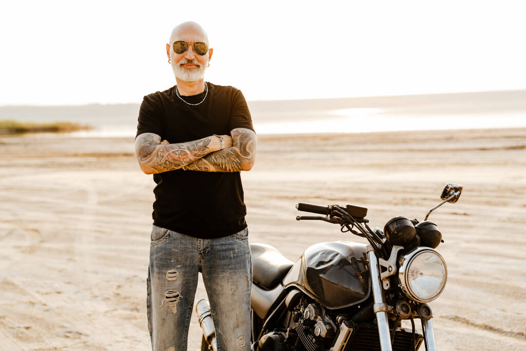 Зрелый смелый мужчина с татуировкой, смотрящий в камеру, позируя на мотоцикле на открытом воздухе - Фото, изображение