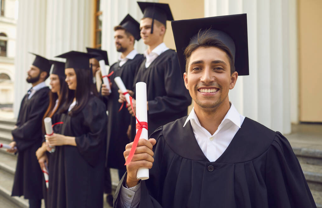 Glücklicher, selbstbewusster Absolvent im Mantel mit einem Diplom in der Hand. - Foto, Bild