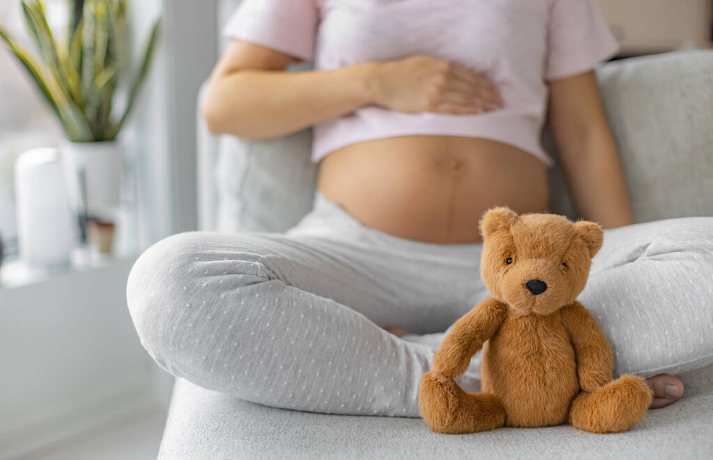 Těhotenství. Těhotná žena na sobě těhotenské oblečení relaxační na domácí pohovce s plyšovým medvídkem dítě hračka v ohnisku pro dítě dárek koncepce nebo očekává dítě - Fotografie, Obrázek