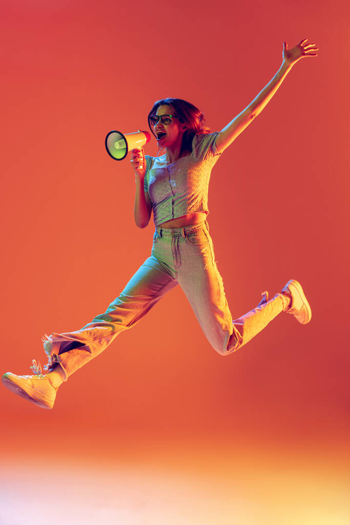 Een slanke mooie opgewonden meisje springen met megafoon geïsoleerd op perzik kleur achtergrond in neon licht, filter. Concept van emoties, muziek, nieuws, info - Foto, afbeelding