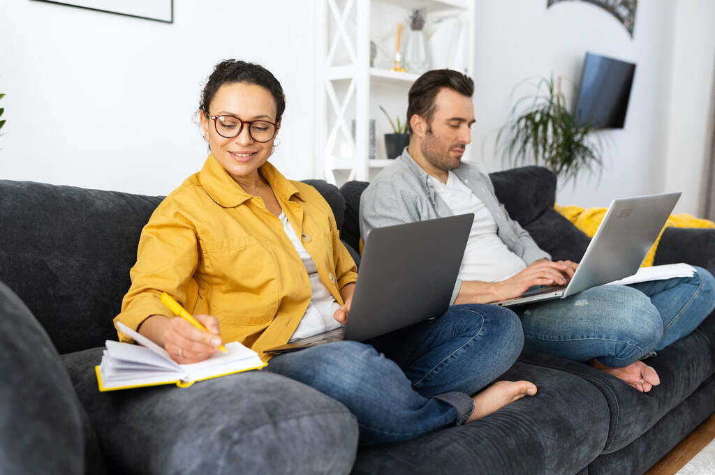 Para korzystająca z laptopów do zdalnej pracy na sofie w domu, skupieni małżonkowie pracujący na odległość, odpowiadający na maile - Zdjęcie, obraz