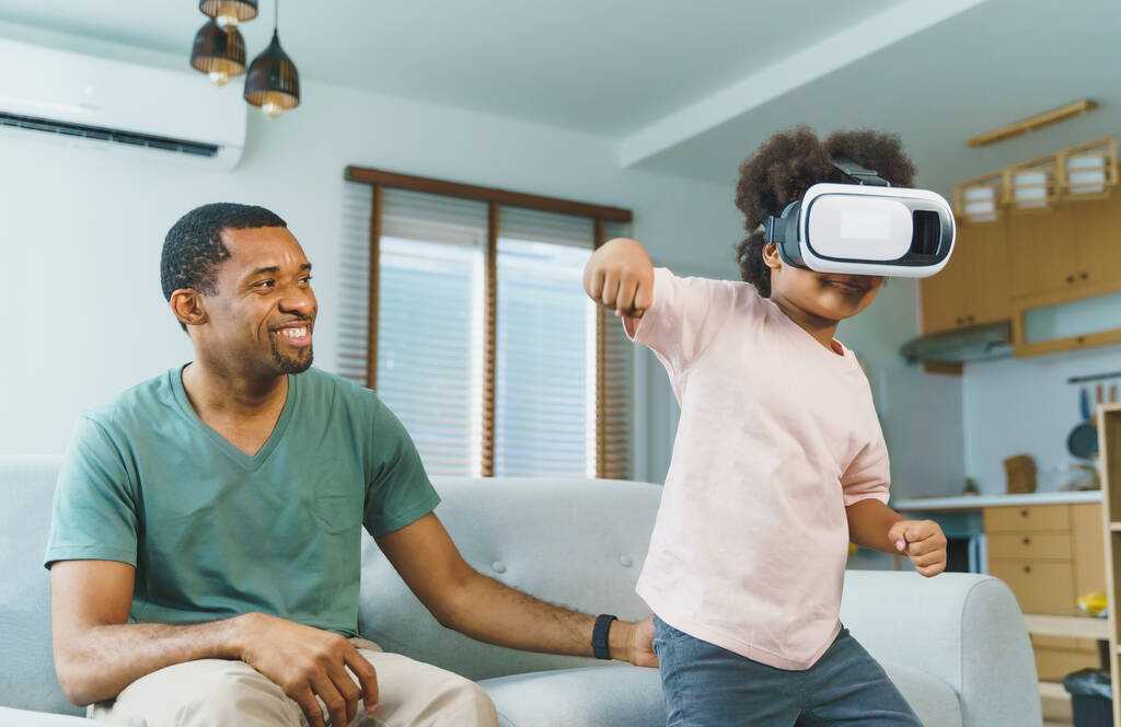 Happy Afro African american boy in VR headset, enjoying playing virtual reality video games with his father at home. - 写真・画像